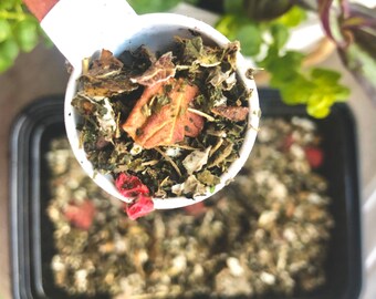 May include: A white measuring spoon filled with a blend of dried herbs and flowers, including green leaves, red berries, and a large, dried, orange-brown leaf. The spoon is held over a black container filled with the same herbal blend.