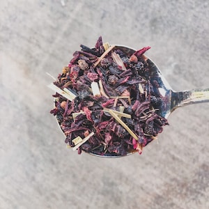 May include: A silver spoon filled with a blend of dried hibiscus flowers, lemongrass, and black peppercorns.