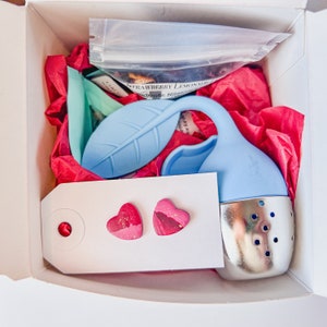May include: A white gift box with pink tissue paper, a blue silicone tea infuser with a leaf design, a small white card with two pink heart-shaped earrings, and a clear plastic bag with loose leaf tea.