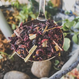 May include: A silver spoon filled with a blend of dried hibiscus flowers, lemongrass, and other herbs. The mixture is a deep red color and appears to be a loose leaf tea.