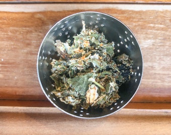 May include: A small, round, stainless steel strainer filled with dried green herbs and spices. The strainer has small holes and sits on a wooden surface. The herbs have a mix of green, brown, and orange hues.