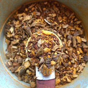 May include: Close-up of a white spoon filled with a blend of dried herbs and spices, including cinnamon sticks, orange peel, and turmeric. The spoon is resting on a bowl of the same blend.