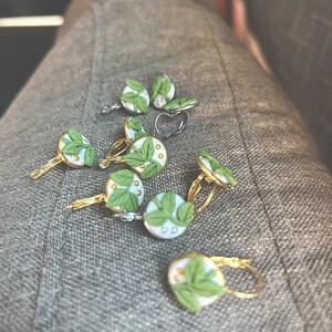 May include: A collection of handmade earrings and a ring with a botanical theme. The earrings feature a gold-colored metal base with a round, white and light blue design, adorned with green leaves and small gold accents. The ring has a silver-colored heart shape.