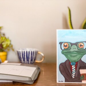 May include: A hand-painted card featuring a cartoon frog wearing glasses and a suit, holding a cup of coffee. The frog has a green body, orange eyes, and a brown suit with a blue and white striped tie. The background is a light blue watercolor wash.