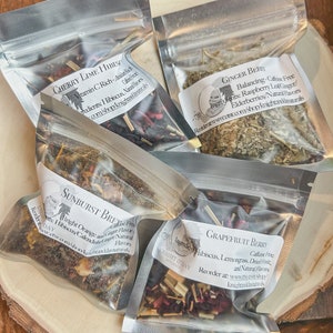 May include: Four small clear plastic bags filled with dried herbs and flowers. The bags are labeled with the names of different herbal tea blends: Cherry Lime Hibiscus, Ginger Berry, Sunburst Brew, and Grapefruit Berry. Each label includes the ingredients and a website address.