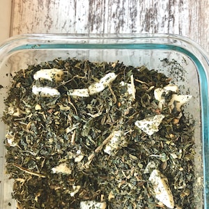 May include: A glass container filled with a mixture of dried green herbs and white pieces of dried fruit. The container is sitting on a wooden surface.