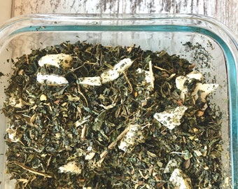May include: A glass container filled with a mixture of dried green herbs and white pieces of dried fruit. The container is sitting on a wooden surface.