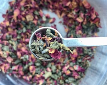 May include: A close-up of a metal teaspoon filled with a blend of dried green tea leaves, rose petals, and lavender buds.