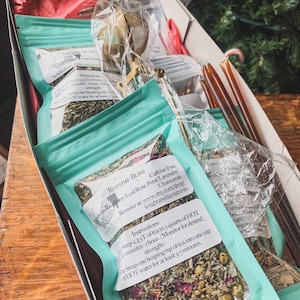 Op de afbeelding: Een turquoise doos gevuld met vier zakjes losse bladthee. De zakjes zijn gelabeld "Bedtime Bliss" en "Caffeine Free Raspberry Leaf/Rose Petal/Lavender/Chamomile". De doos bevat ook een gouden thee-infuser en twee houten honingroerstaafjes.