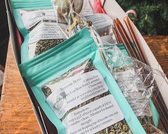 Könnte beinhalten: Eine türkisfarbene Box gefüllt mit vier Beuteln losen Blatttees. Die Beutel sind mit "Bedtime Bliss" und "Caffeine Free Raspberry Leaf/Rose Petal/Lavender/Chamomile" beschriftet. Die Box enthält außerdem einen goldenen Tee-Aufguss und zwei hölzerne Honigrührer.