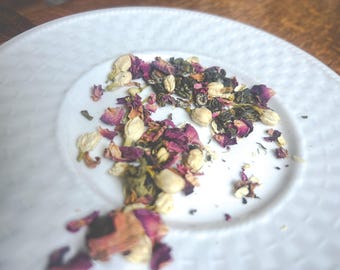 May include: A white plate with a mixture of dried green tea leaves, rose petals, and jasmine flowers.