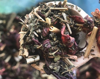 May include: Close-up of a wooden spoon filled with a blend of dried herbs and flowers, including green leaves, red hibiscus petals, and thin pieces of dried lemongrass.