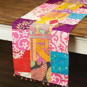 May include: A colorful patchwork table runner with a variety of patterns and colors, including pink, purple, yellow, and blue. The runner is made of stitched fabric and has small tassels at the ends. It is displayed on a wooden table.
