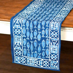 May include: A rectangular fabric table runner featuring a deep blue background with a repeating pattern of light blue fish. The runner is bordered by a white geometric design consisting of squares, triangles, and intersecting lines on a blue base.