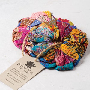 Puede incluir: Un scrunchie para el pelo de tela colorida hecho de saris reciclados. El scrunchie está hecho por mujeres en la India utilizando la técnica Kantha Craft. La etiqueta dice "SÉVYA HANDMADE This product is made from up-cycled saris by women in India using the Kantha Craft Sevya is a member of the Fair Trade Federation www.sevya.com"