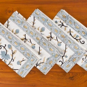 May include: Four rectangular cloth napkins arranged in a row, each featuring a white base with a repeating floral pattern in muted blue, yellow, and black. The design includes a wide decorative border with scrolling vines and central floral motifs.