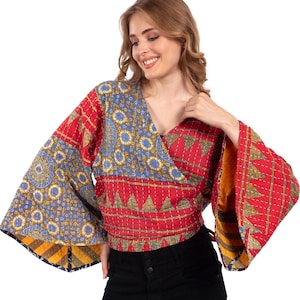 Op de afbeelding: Een wikkel top met lange, uitlopende mouwen. De top heeft een patchwork ontwerp met rode, blauwe en gele patronen. De stof lijkt gewatteerd te zijn, met zichtbare stiksels. De top wordt in de taille vastgebonden.