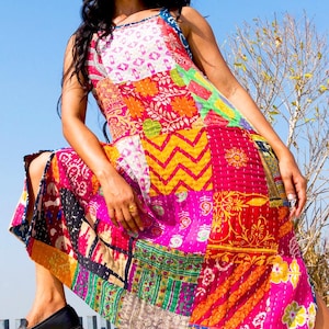 May include: A woman wearing a colourful patchwork maxi dress with a variety of floral and geometric patterns. The dress is sleeveless and has a relaxed fit. The woman is standing on a white surface with a blue sky in the background.