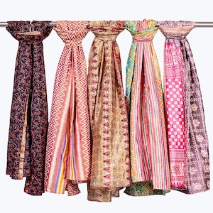 Könnte beinhalten: Fünf lange Schals hängen an einer horizontalen Metallstange. Die Schals weisen verschiedene Muster auf, darunter florale, Zickzack-, Paisley- und geometrische Designs in Farben wie Marineblau, Pink, Gold, Grün und Violett mit feinen Details.