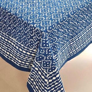 May include: A blue and white patterned tablecloth. The tablecloth features a repeating white floral design on a blue background. The border has a white wavy stripe design. The tablecloth is made of a soft, durable fabric.