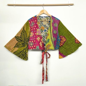 Puede incluir: Un top corto cruzado con mangas largas y anchas, con un diseño de patchwork que combina varios patrones florales y geométricos en tonos verde, rosa, amarillo y azul. La prenda está confeccionada con costuras visibles de estilo kantha.