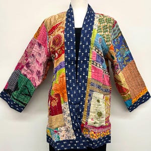 May include: A quilted jacket featuring a patchwork design of various colorful fabrics in shades of pink, orange, green, and blue. The garment has a dark blue collar and trim with a small white floral pattern, long sleeves, and a straight hemline.