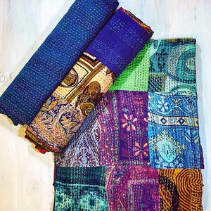 May include: A folded textile featuring a patchwork design with various patterns in shades of blue, purple, green, and yellow. The fabric displays intricate kantha stitching throughout, creating a textured surface with visible thread lines across the quilt.