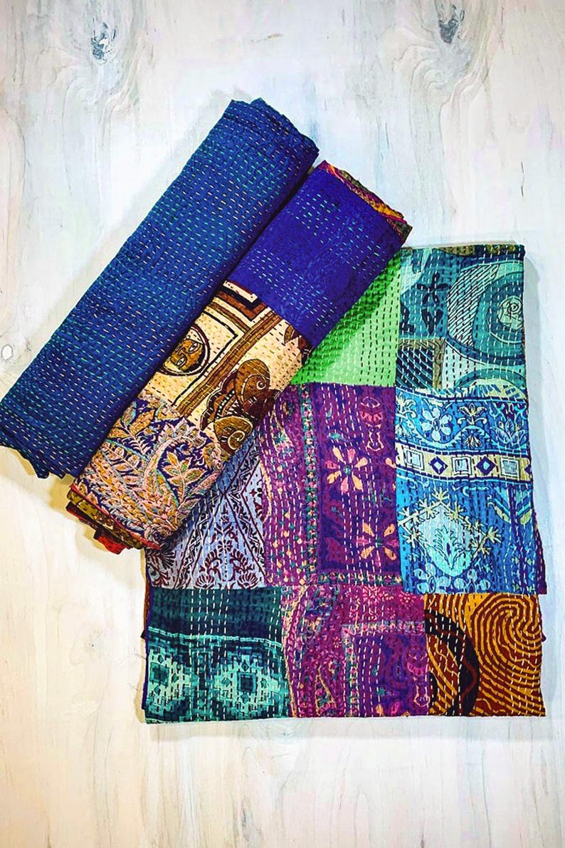 May include: A folded textile featuring a patchwork design with various patterns in shades of blue, purple, green, and yellow. The fabric displays intricate kantha stitching throughout, creating a textured surface with visible thread lines across the quilt.