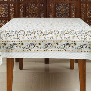 May include: A white tablecloth with a floral pattern and a blue and gold border. The tablecloth is on a wooden table with four legs.