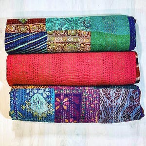 May include: Three folded quilts stacked vertically. The top quilt features a patchwork of teal, blue, and floral patterns. The middle quilt is solid bright pink with visible yellow stitching. The bottom quilt displays a patchwork of blue, purple, and floral designs.