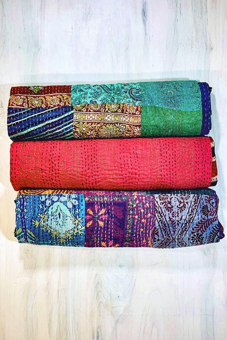 May include: Three folded quilts stacked vertically. The top quilt features a patchwork of teal, blue, and floral patterns. The middle quilt is solid bright pink with visible yellow stitching. The bottom quilt displays a patchwork of blue, purple, and floral designs.