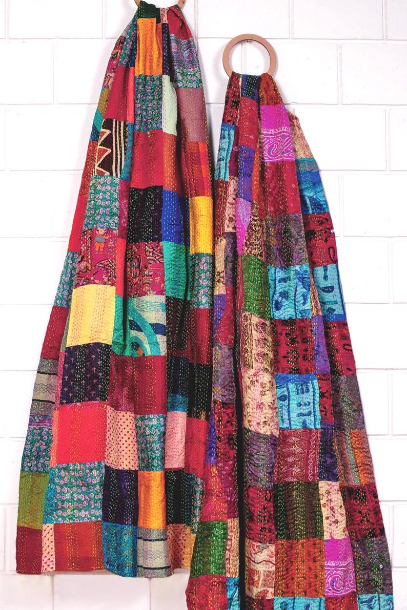 May include: Two baby carriers made from multicoloured patchwork fabric featuring various patterns and textures. Each carrier is suspended from a circular wooden ring at the top, showcasing a vibrant array of red, blue, yellow, and pink squares.