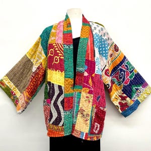 May include: A patchwork jacket featuring a variety of colorful fabric panels with diverse patterns including floral, geometric, and abstract designs. The garment has long sleeves, an open front, and visible white stitching throughout the fabric.
