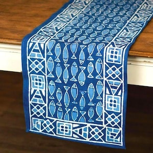 May include: A blue table runner with a white fish pattern. The runner has a repeating design of fish and geometric shapes. The runner is made of fabric and is placed on a wooden table.