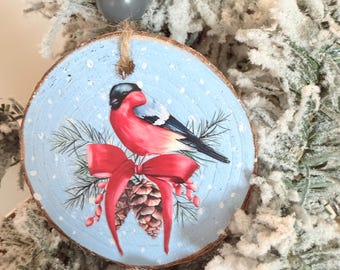 May include: A round wooden Christmas ornament featuring a painted bird with red and black feathers, a red bow, and pine cones on a light blue background. The ornament is suspended by a twine loop and a gray bead.