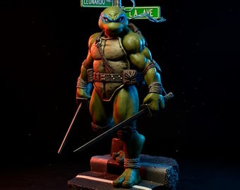 May include: A green Leonardo Teenage Mutant Ninja Turtle statue holding two katanas, standing on a street-themed base. The statue has a blue mask and is posed in a dynamic stance. Street signs read "Leonardo" and "L.A. Ave."