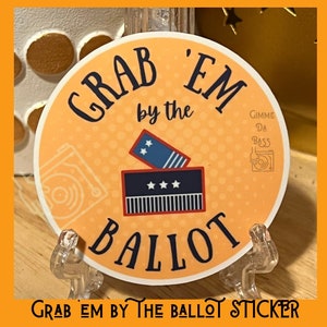 May include: A round sticker with a yellow background and the text "Grab 'Em by the Ballot" in black. The sticker features a red, white, and blue ballot box with stars and stripes. The text "Gimme Da Bass" is in a small white box in the upper right corner.