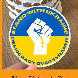 May include: A round sticker with a blue and yellow background, a white fist raised in the air, and the text "Stand with Ukraine Democracy Over Tyranny" in a white circle.