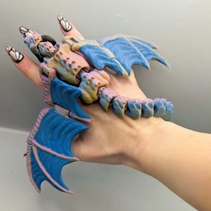 May include: A multi-colored articulated dragon claw, featuring blue, pink, and yellow hues. The claw is designed to be worn on the hand, with a segmented body and wing-like structures. The dragon claw is a unique accessory.