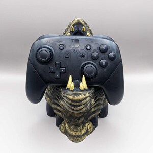 May include: A black Nintendo Switch Pro controller rests on a detailed, gold and brown dragon head-shaped stand. The controller features buttons, joysticks, and the Nintendo Switch logo. The stand has sharp teeth.