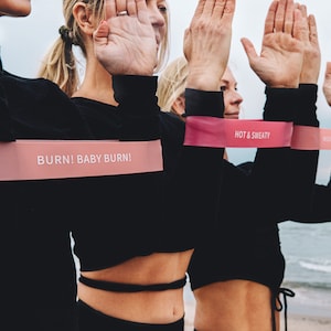 Puede incluir: Cuatro mujeres con camisas negras de manga larga y bandas de resistencia rosas alrededor de sus brazos. Las bandas tienen el texto "BURN! BABY BURN!"  "HOT & SWEATY" y "MOVE WITH PURPOSE" impreso en ellas. Las mujeres están de pie en una playa con el océano de fondo.