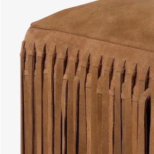 May include: Close-up of a brown suede ottoman with a fringed skirt. The ottoman has a rounded top and a fringe that hangs down from the base. The color is a warm, earthy brown.
