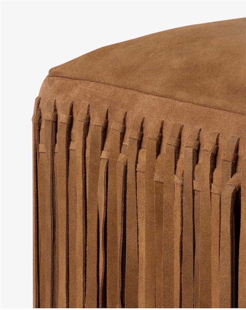 May include: Close-up of a brown suede ottoman with a fringed skirt. The ottoman has a rounded top and a fringe that hangs down from the base. The color is a warm, earthy brown.