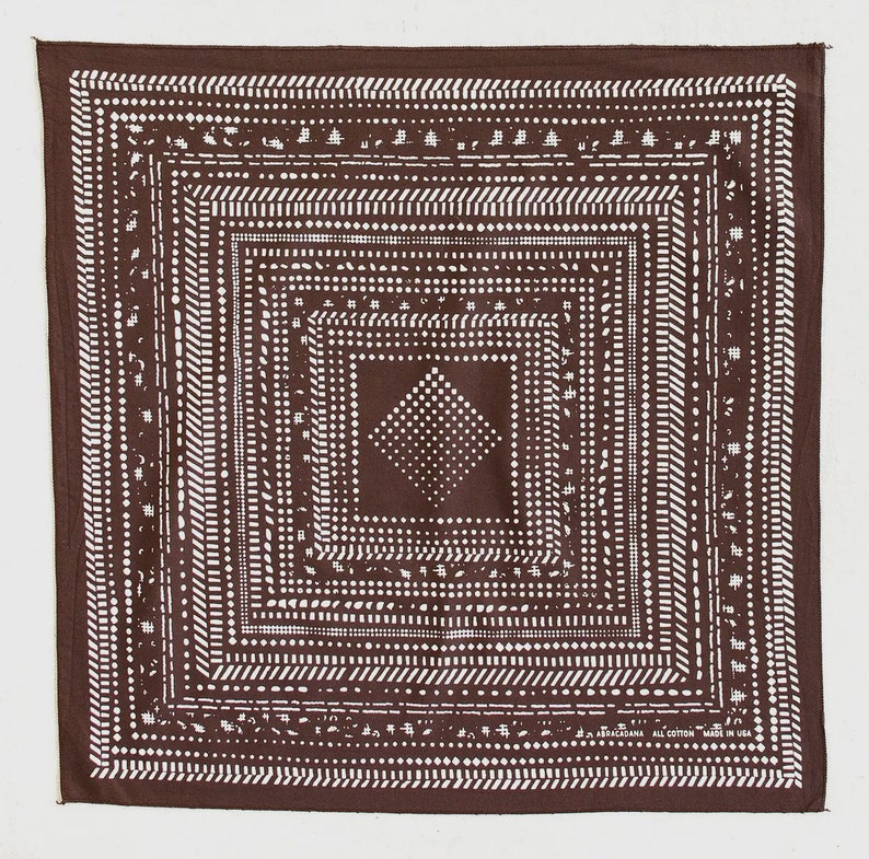 May include: A square, brown bandana with a white geometric pattern. The design features concentric squares and a central diamond shape, all composed of dots and lines. The bandana has a border with the text "ABRACADANA ALL COTTON MADE IN USA".