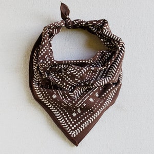 May include: A brown bandana with a white geometric pattern. The bandana is knotted at the top and has a square shape. The pattern includes dots, lines, and small hash marks. The fabric appears to be soft and the overall design is simple and stylish.