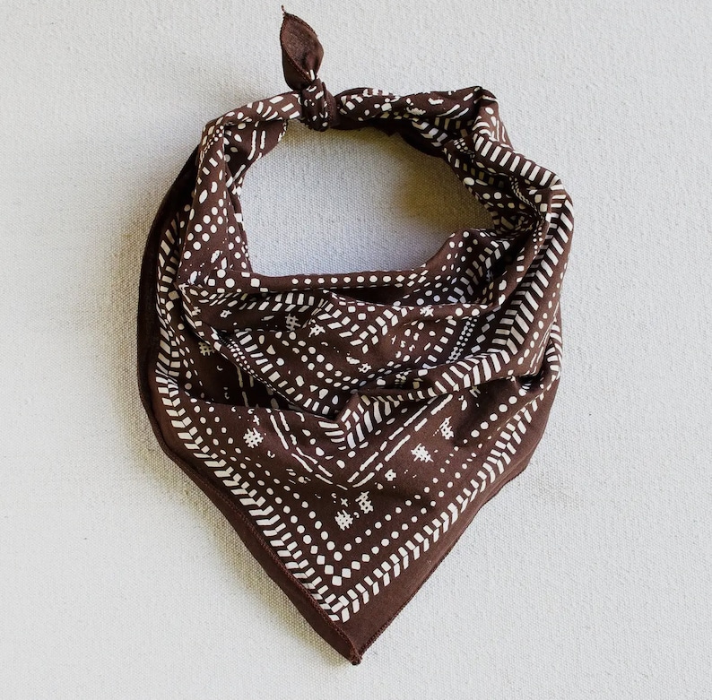 May include: A brown bandana with a white geometric pattern. The bandana is knotted at the top and has a square shape. The pattern includes dots, lines, and small hash marks. The fabric appears to be soft and the overall design is simple and stylish.