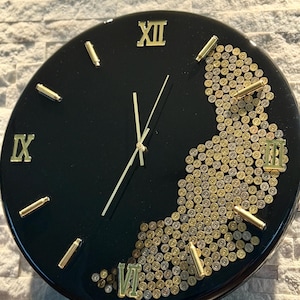 May include: Black round wall clock with gold Roman numerals and hands. The clock face is decorated with gold bullet casings. The clock has a glossy finish and is mounted on a light-colored textured wall.