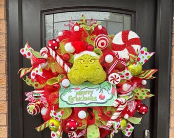 May include: A decorative wreath featuring a plush green character head wearing a red Santa hat, surrounded by red and lime green mesh, candy cane ornaments, peppermint swirls, and a sign that reads Merry Grinchmas in green and red text.