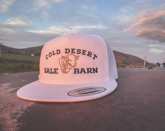 Może przedstawiać: Biała czapka trucker z haftowanym logo w kolorze czarnym i brązowym, na którym widnieje napis "Cold Desert Sale Barn" oraz głowa byka w środku.
