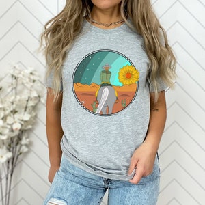 Op de afbeelding: Een grijs T-shirt met een afbeelding van een persoon die op een paard rijdt in een woestijnlandschap met een grote gele bloem en een sterrenhemel.